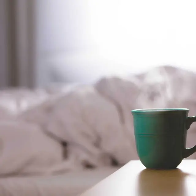 Cama con sábanas blancas y una taza verde sobre la mesilla