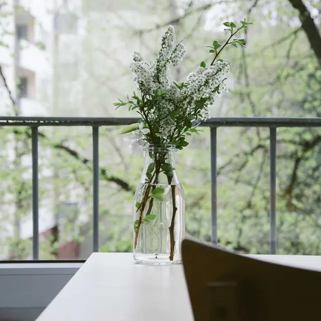 Jarrón con flores sobre una mesa frente a un balcón con árboles